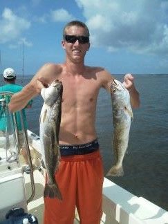 Man on a boat holding two large fish, with blue ocean and sky in the background.