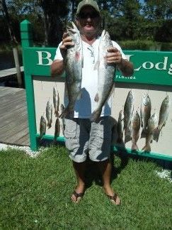 Person holding two fish in front of a fish-cleaning station with more fish displayed on the sign.