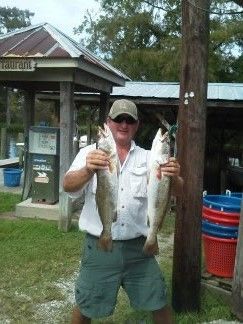 Man holding two fish in front of a park shelter, wearing sunglasses and a cap.