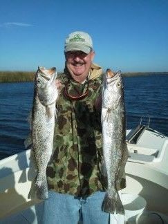 Person on a boat holding two large fish over the water, wearing a camouflage shirt and cap.