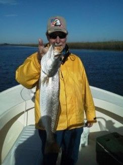 Person on a boat holding a large fish, wearing a yellow jacket and cap on a sunny day.