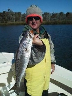 Person in yellow waders and sunglasses holding a large fish on a boat with water and trees in the background