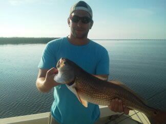 Man holding a large fish by a calm lakeshore at sunset, wearing a blue shirt and sunglasses