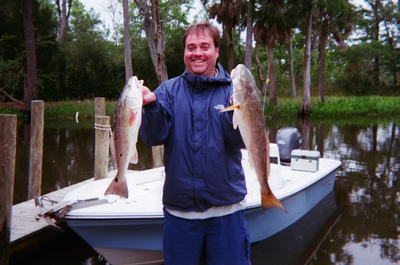 Person smiling and holding two large fish in front of a boat on a wooded canal