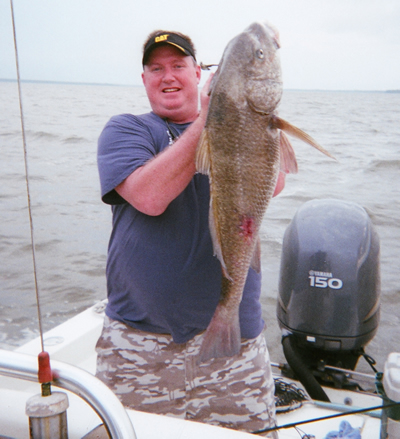 Man holding a large fish on a boat, with calm water and an outboard motor in the background