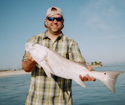 Man holding a large silver fish on a beach, wearing a plaid shirt, cap, and sunglasses.