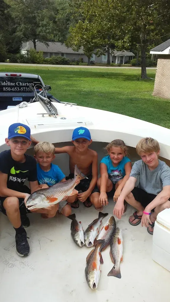 Five kids crouch on a boat deck holding several large fish, with a grassy shoreline behind them.