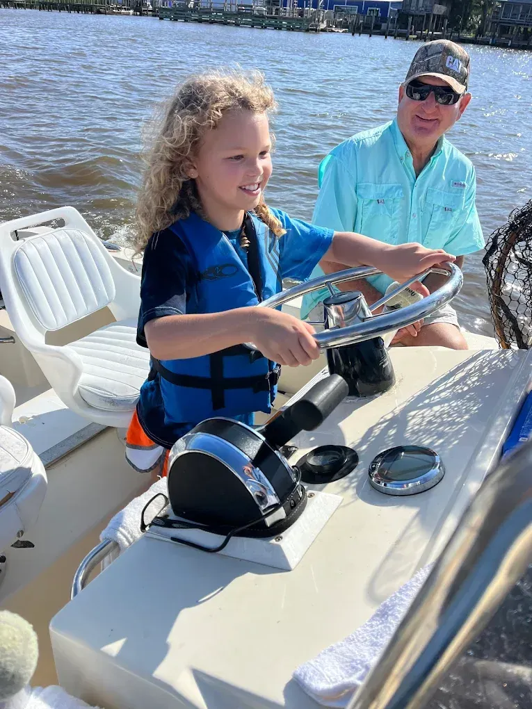 Child steering a boat on the water with an adult seated behind them on a sunny day