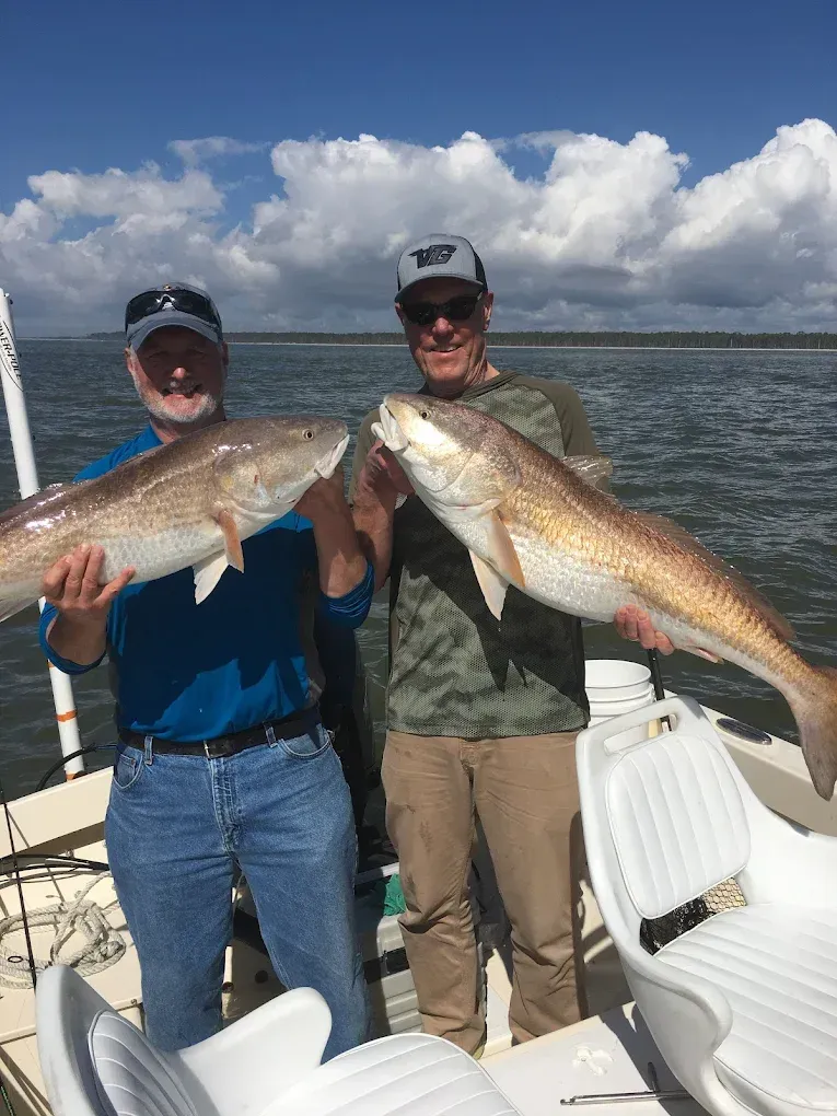 Two people on a boat holding large fish, with calm water and cloudy sky behind them.