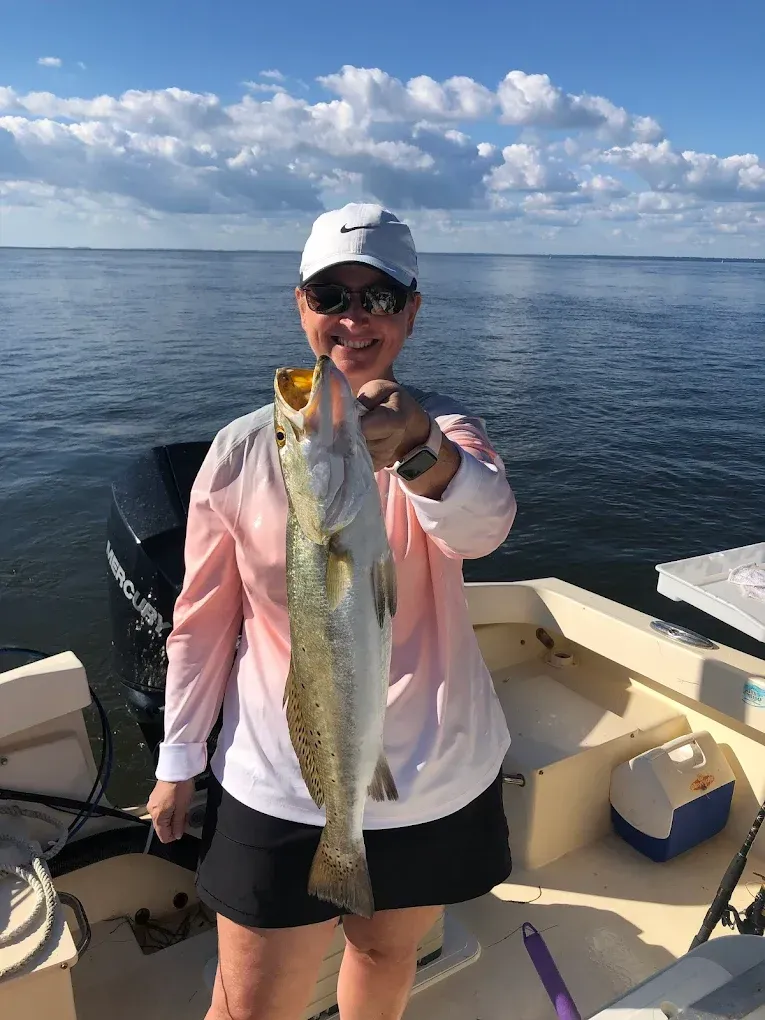 Person on a boat holding a long fish, wearing a pink shirt and white cap on calm water under a cloudy sky
