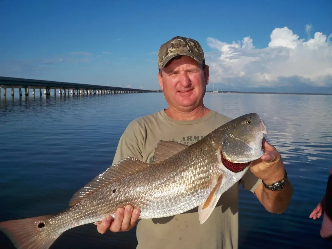 Man holding a large fish on a dock with calm water and a bridge in the background