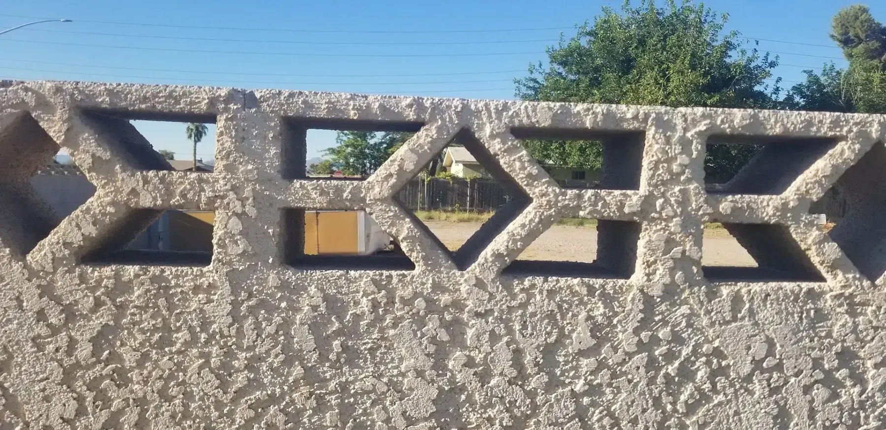 Gray concrete fence with diamond and x-shaped cutouts, with greenery and blue sky in the background.