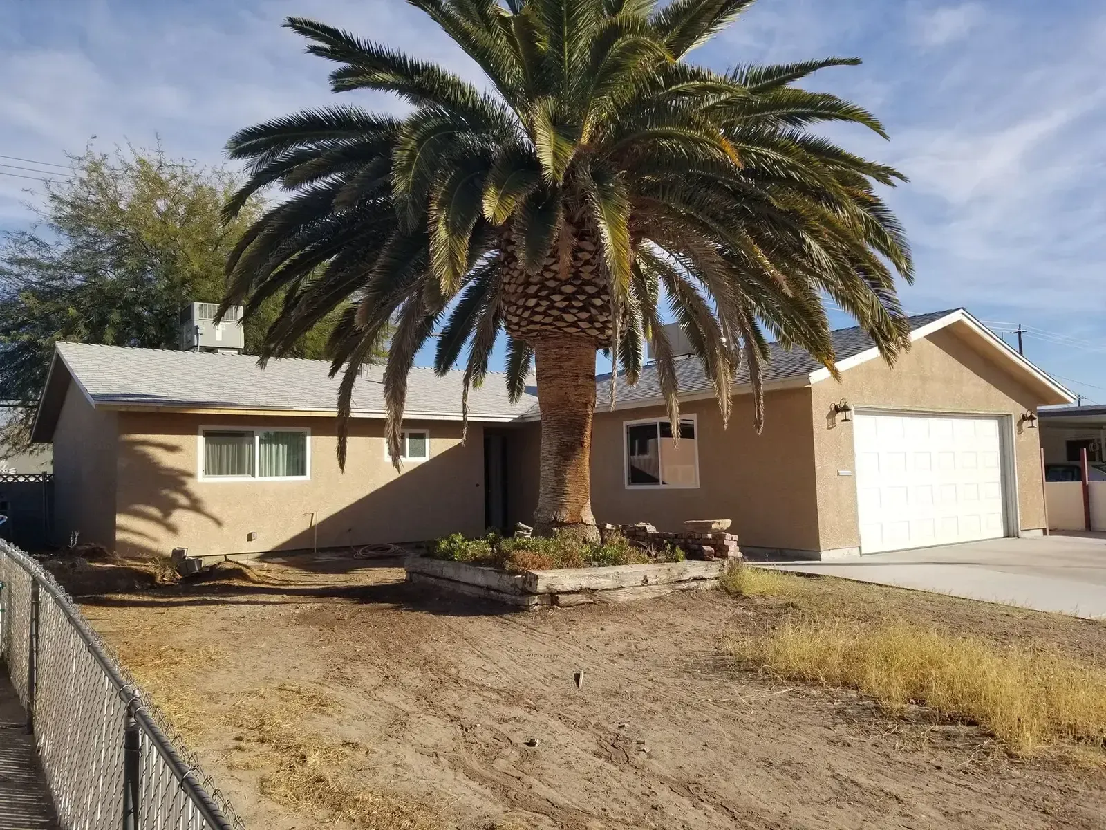 Tan stucco house with palm tree in front yard, dry grass, and a white garage door.