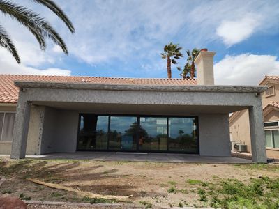 Covered patio with sliding glass doors, stucco walls, and a chimney against a blue sky with palm trees.