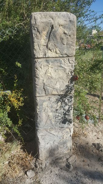 Stone pillar, rectangular shape, in a grassy area, next to a chain-link fence and foliage.