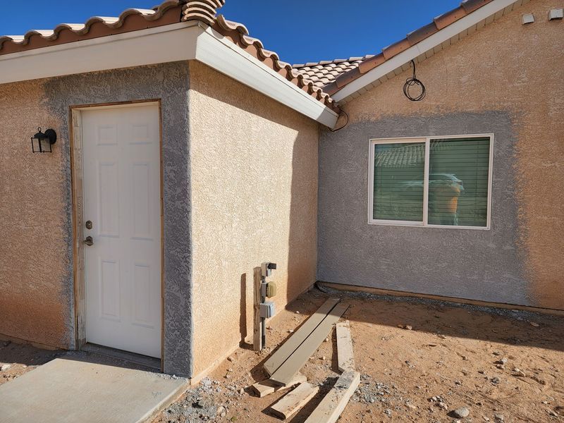 Exterior of a stucco house with a white door and window, tan and gray tones, unfinished ground.