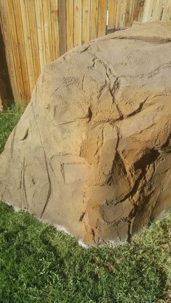 Large, textured rock in front of a wooden fence on green grass.