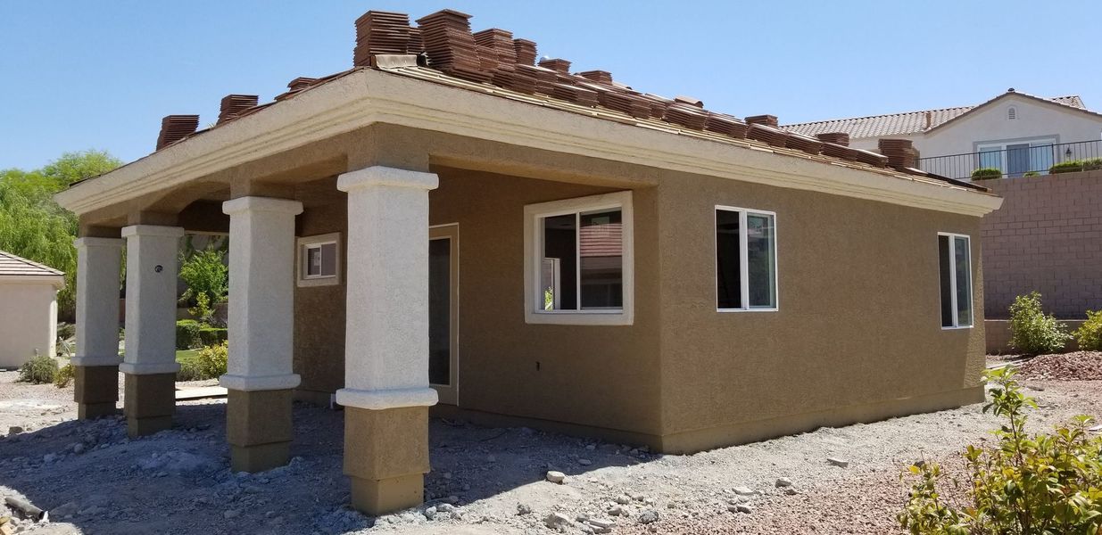 A small, tan stucco house with white pillars and a red tile roof, set on a gravel lot under a bright blue sky.