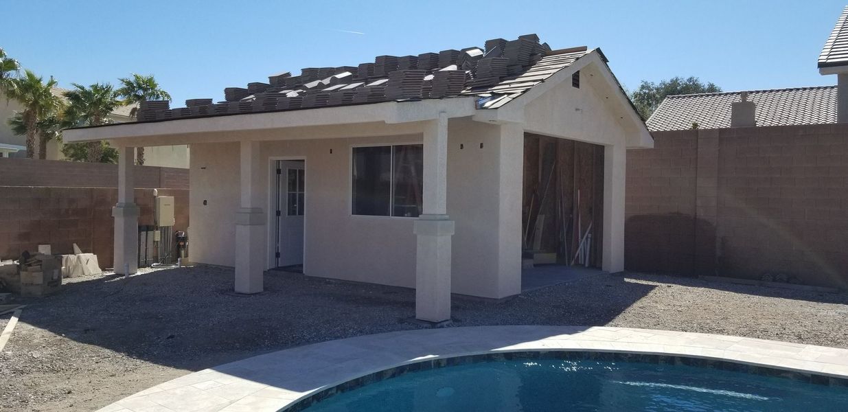 Poolside building with partially finished roof and open garage door, gravel ground, blue sky.