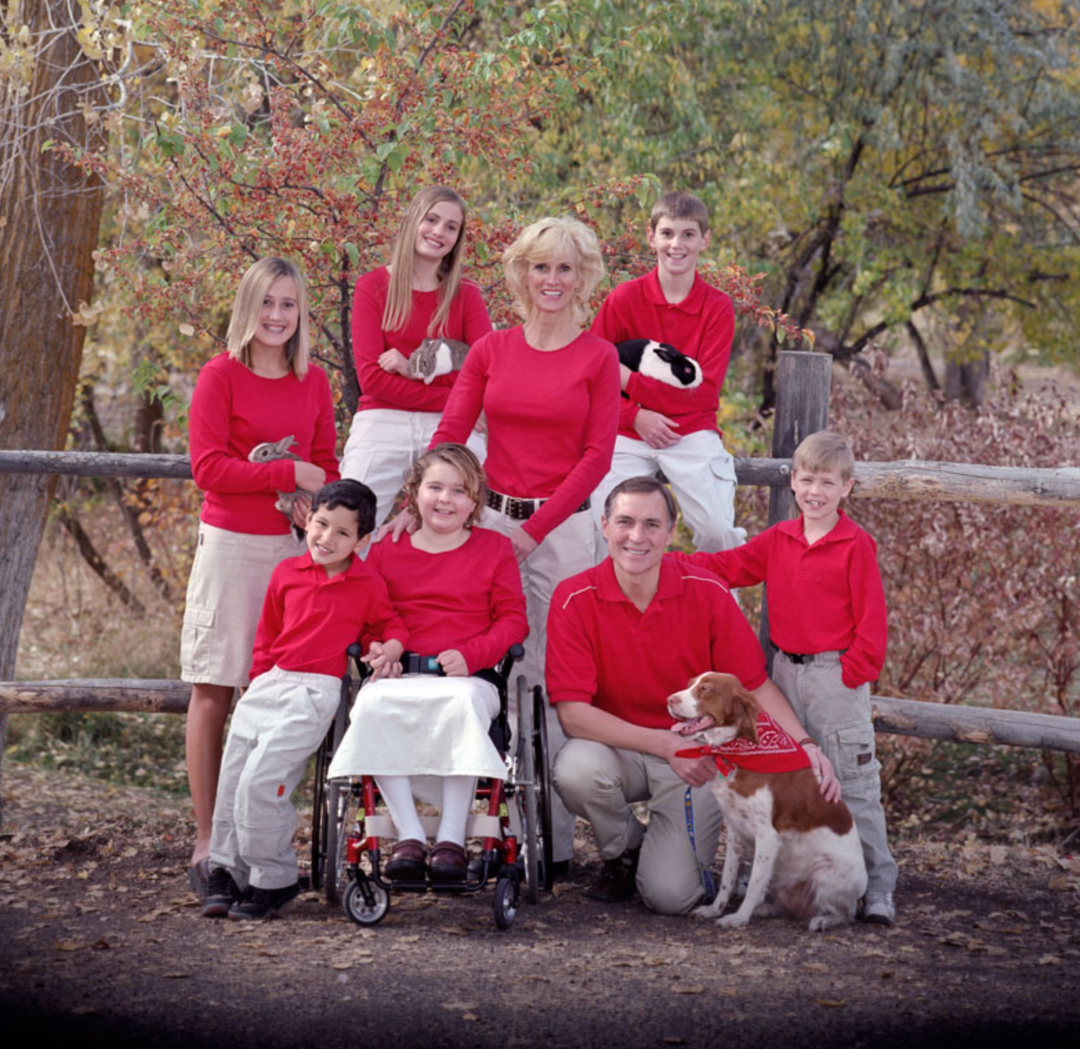 Family in red sweaters posing outdoors in a wooded area with a child in a wheelchair.