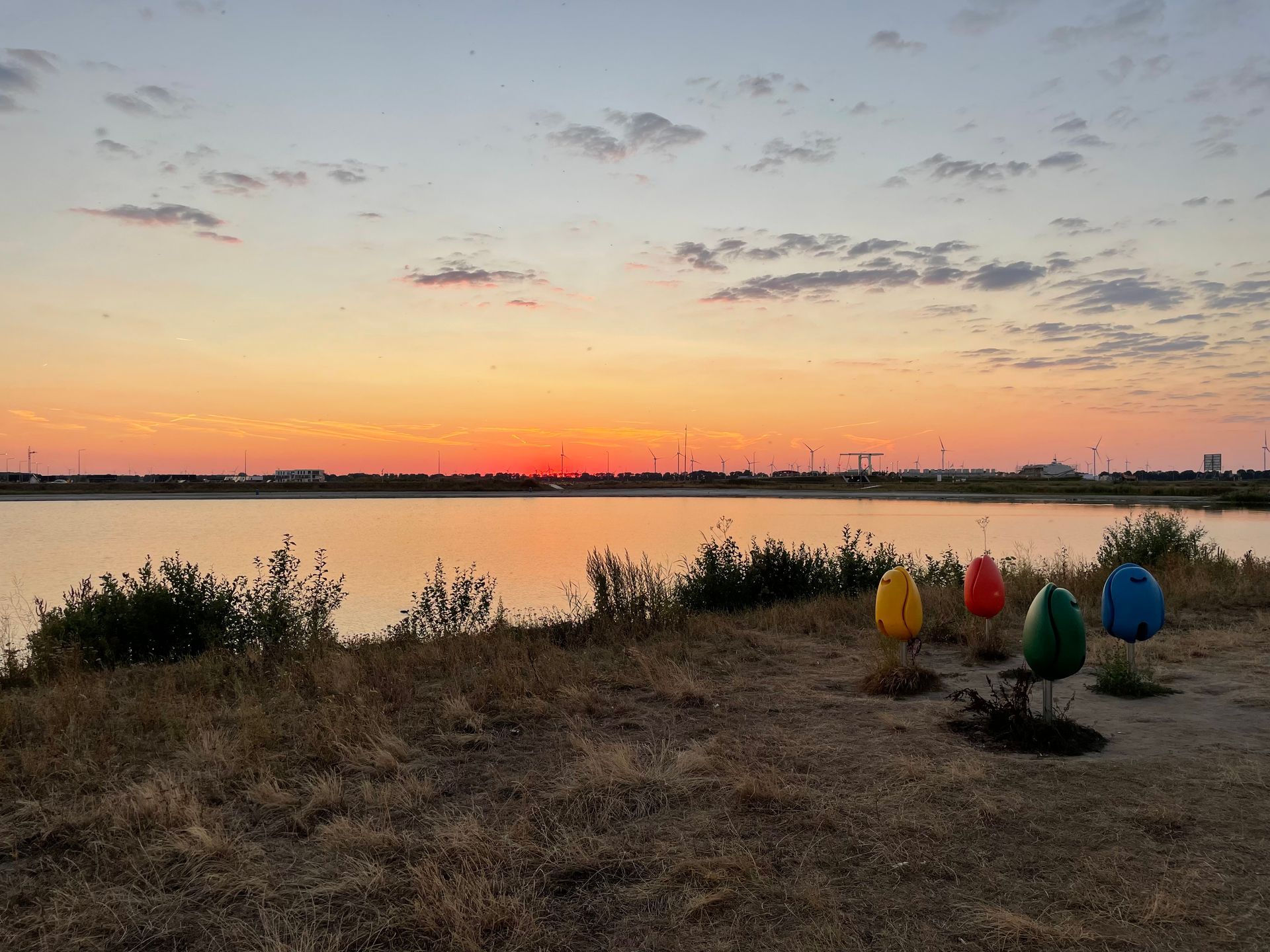 Zonsondergang boven een kalm meer, kleurrijke kunstinstallaties aan de oever, gele, rode, turquoise en blauwe eieren.