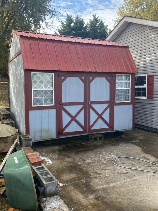 A barn shed with a red roof is in a driveway next to a house.