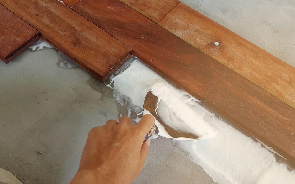 A Person Is Using A Spatula To Spread White Paint On A Wooden Floor — Affordable Carpets Woolgoolga In Woolgoolga, NSW