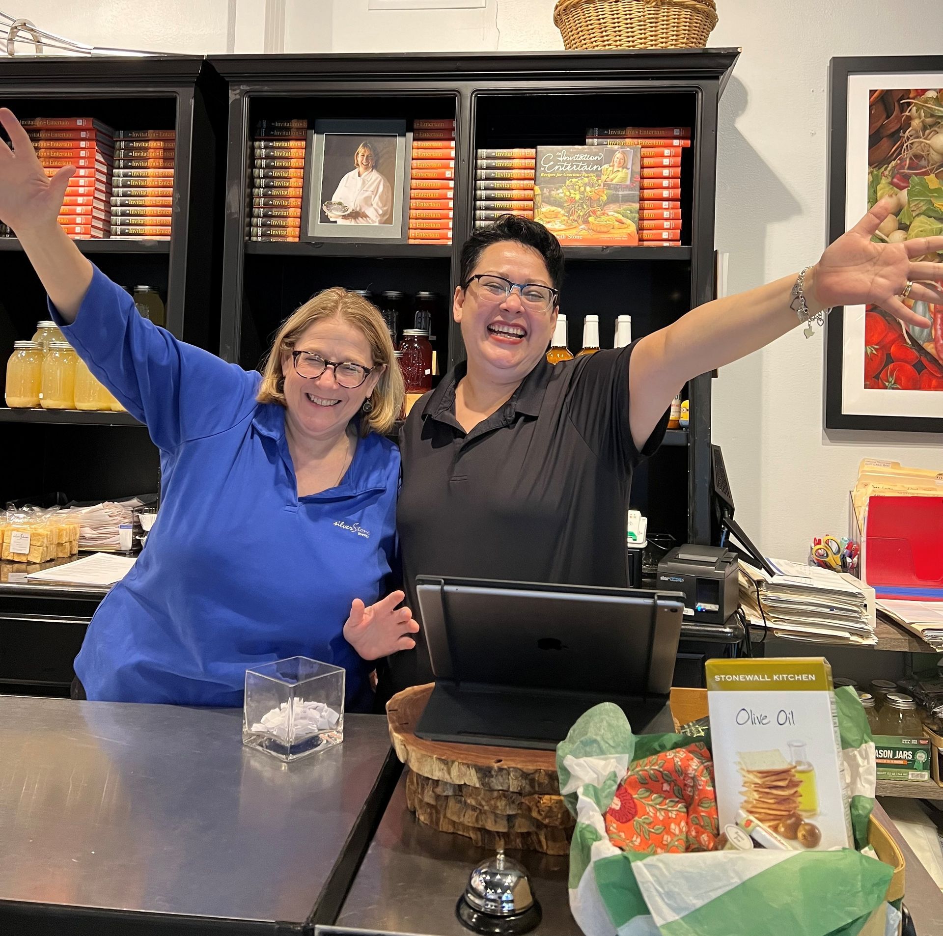 Two people with arms raised behind a counter in a shop; shelves with products in background.