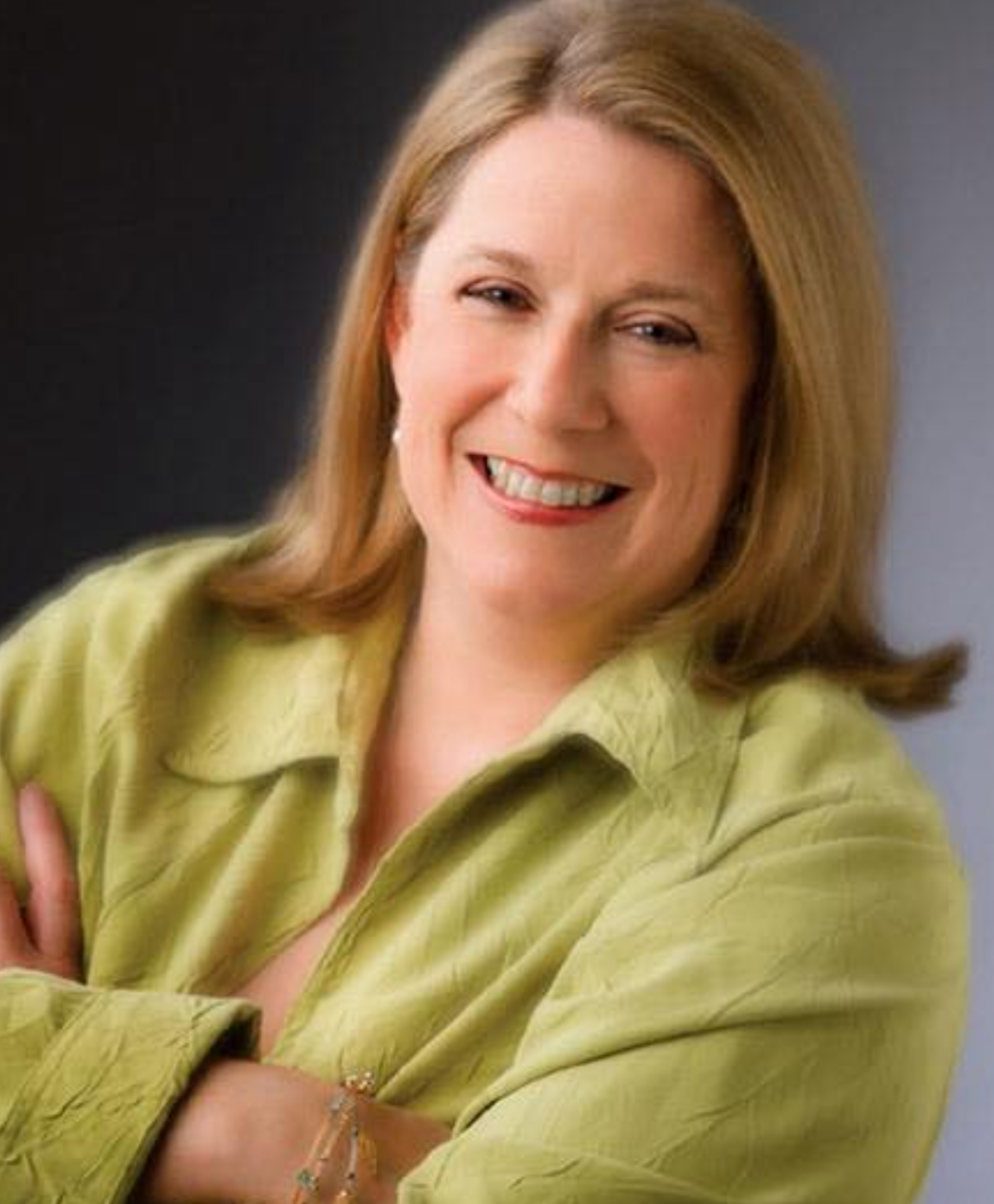 Woman in green blouse smiles, arms crossed, studio backdrop.