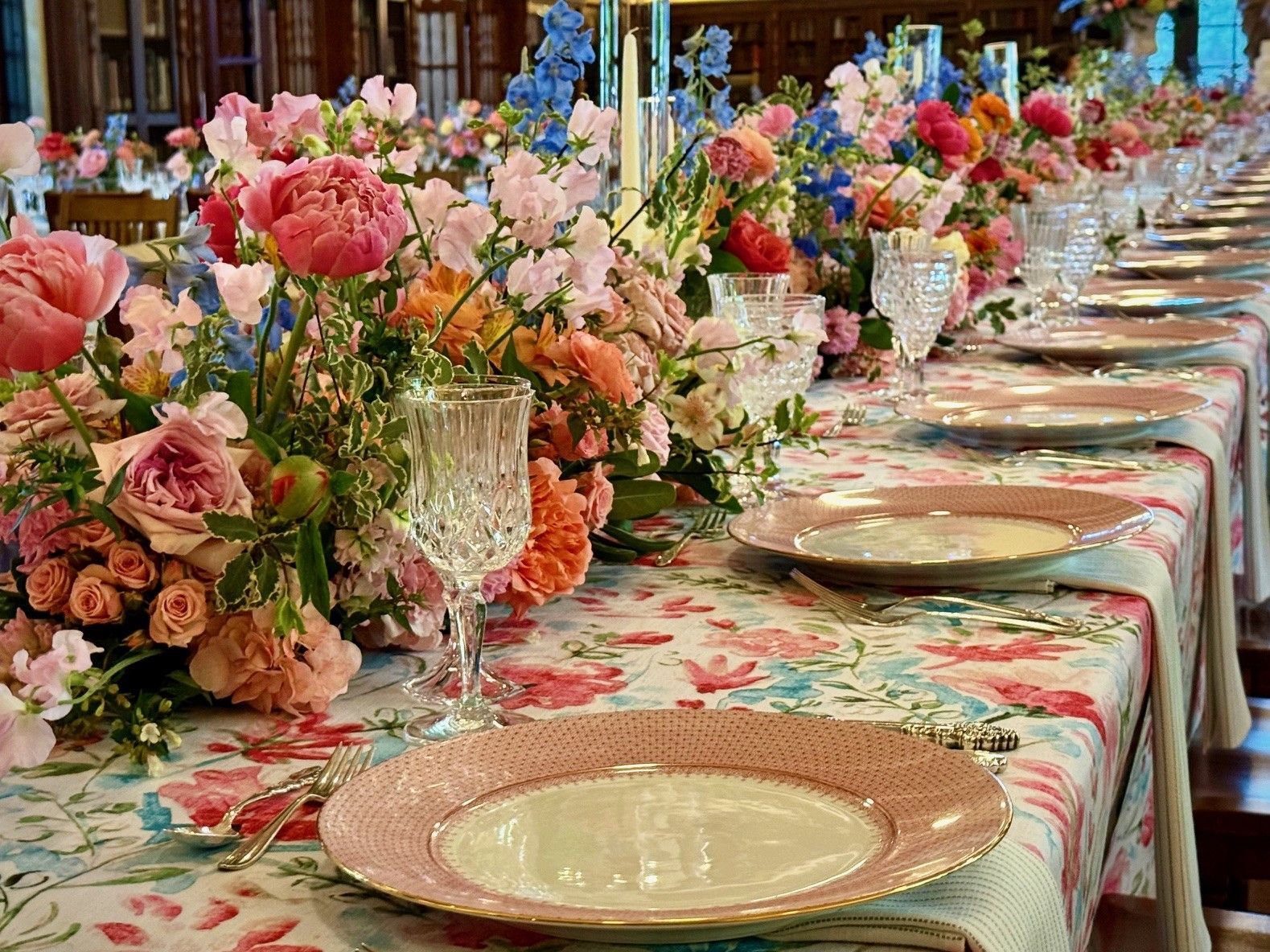 Long table set for a meal, floral centerpiece with pink and blue flowers, pink plates.