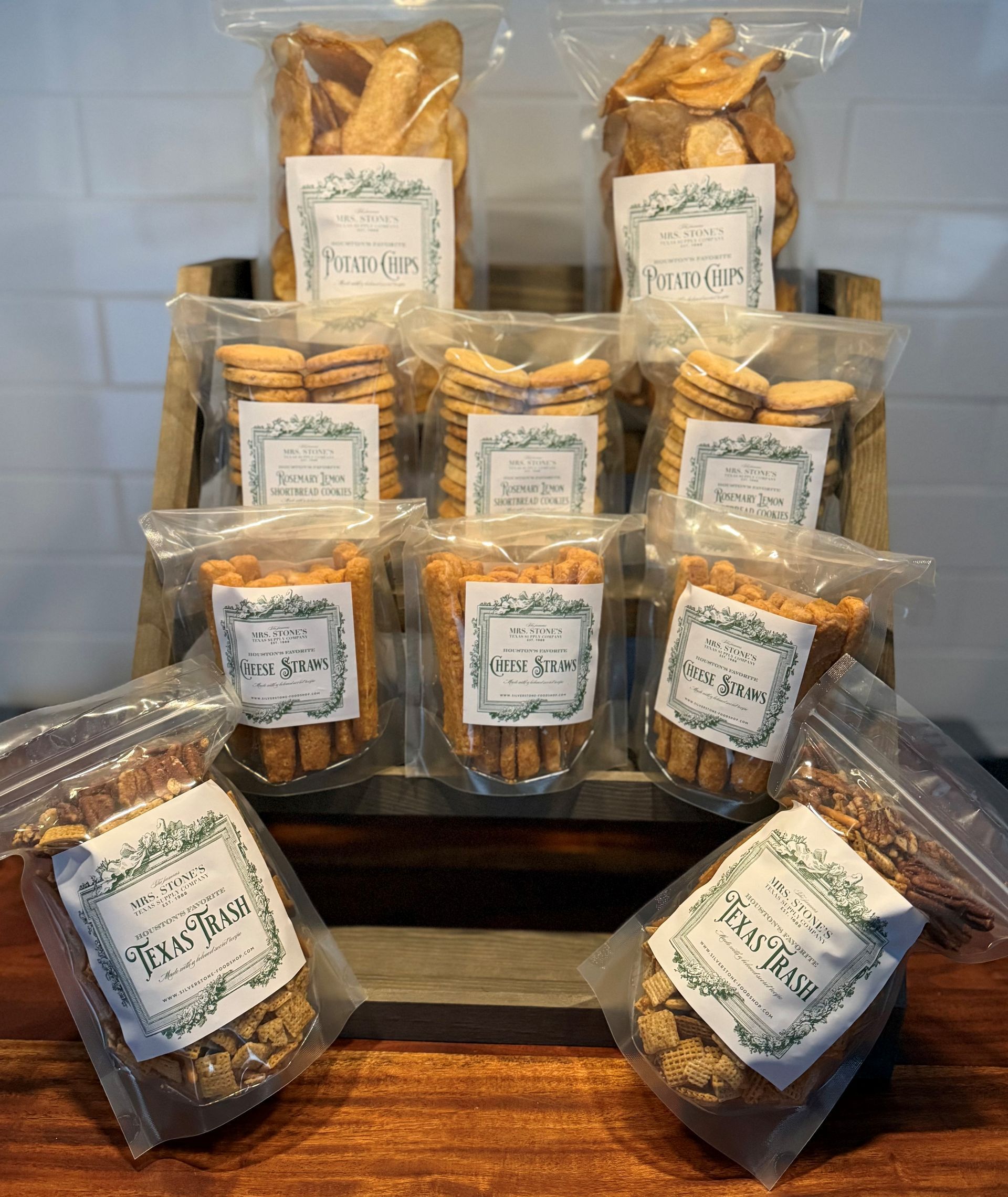 Bags of various baked goods displayed on a wooden stand against a white brick wall.