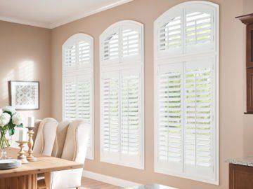 A dining room with a table and chairs and white shutters on the windows.