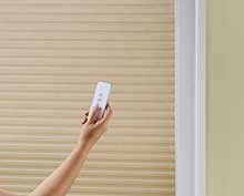a person is holding a remote control in front of a window cellular shade blind.