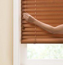 a woman is opening a standard cordlock /cord tilt wooden blind on a window .