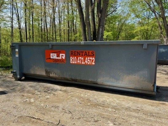 A large, grey metal dumpster sits on a dirt surface in front of a forest, marked with 