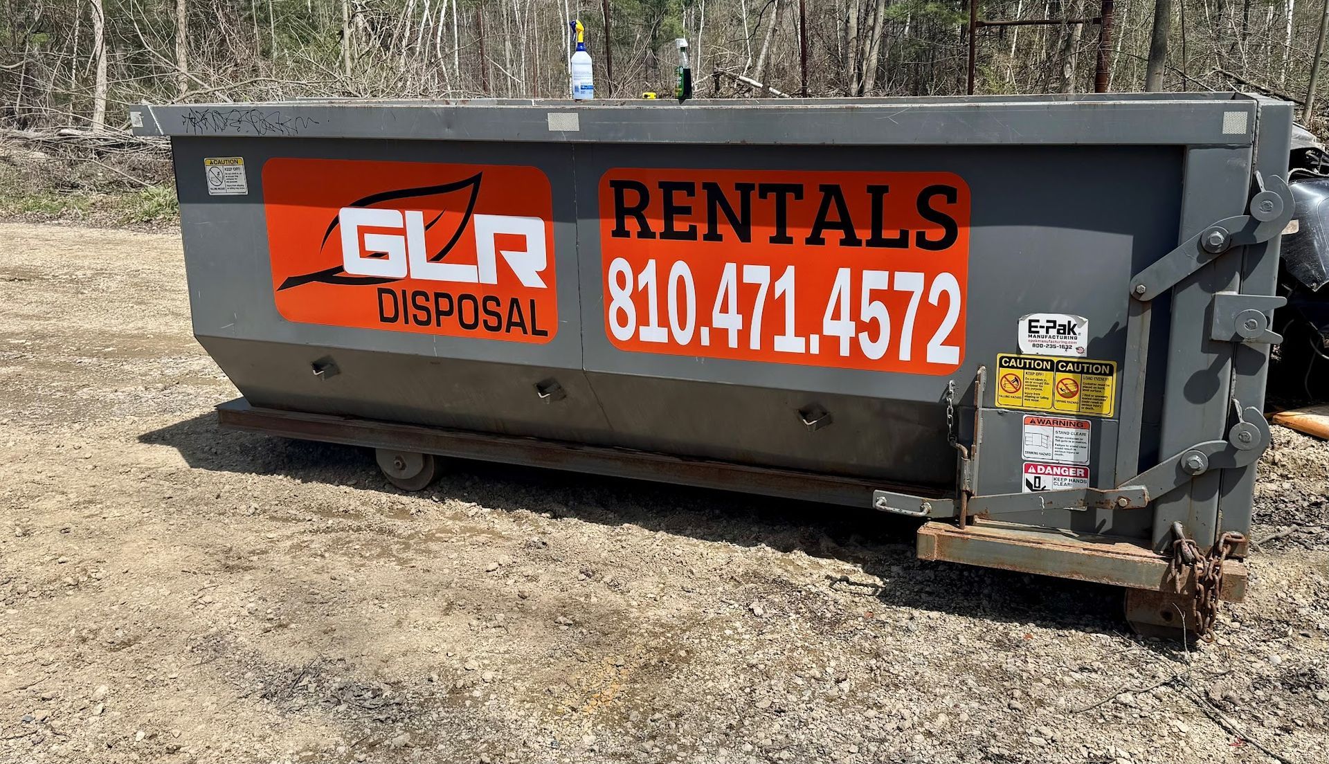 A gray dumpster for GLR Disposal sits on a dirt lot, featuring orange signage with contact information and rentals.