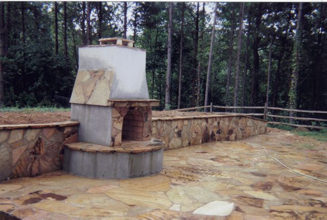 A stone fireplace in the middle of a patio with trees in the background.
