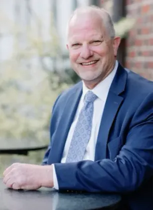 Smiling older man in blue suit, seated outdoors by a brick wall.