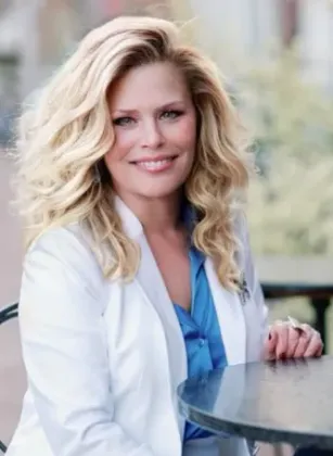 Blonde woman in white jacket smiles, sitting outdoors.