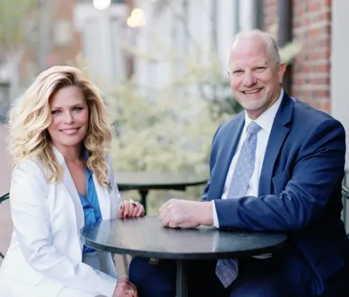 Woman and man sit at a table outside. The woman wears white, the man a blue suit; both smile.