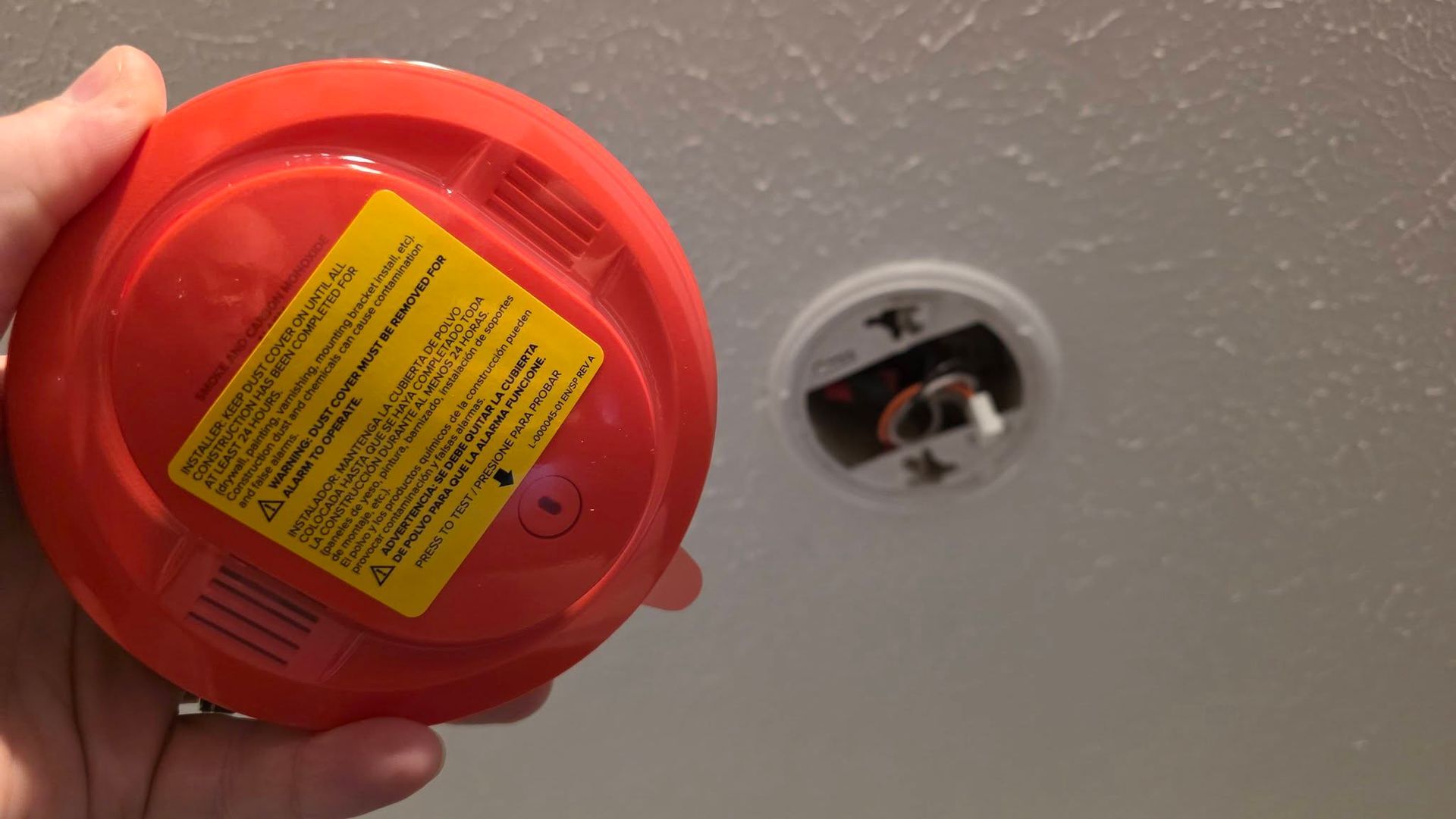 Hand holding a red smoke detector near a ceiling smoke alarm mount with exposed wires