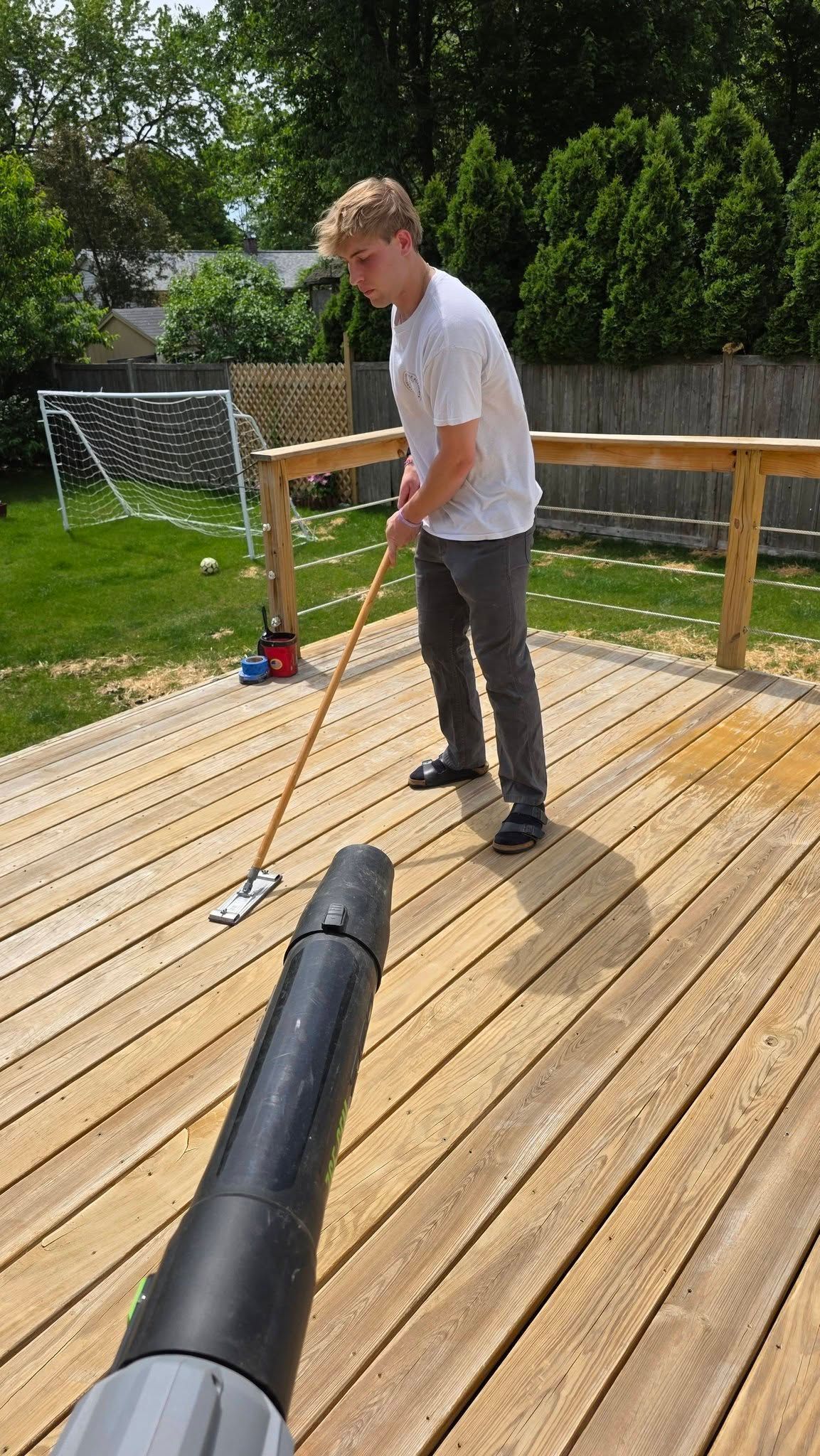 Person using a long pole on a sunny wooden deck with a yard and trees in the background