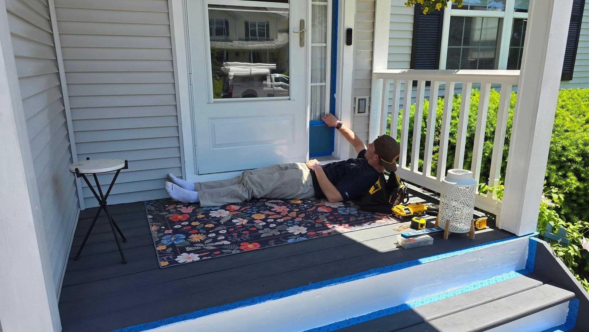 Person lying on a front porch, relaxing on a colorful rug near a white railing and blue-trimmed steps.