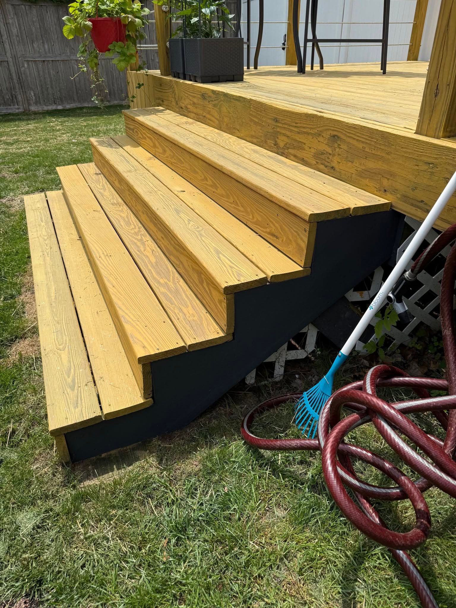 Wooden deck stairs with black skirting beside a grassy yard, with a red hose nearby