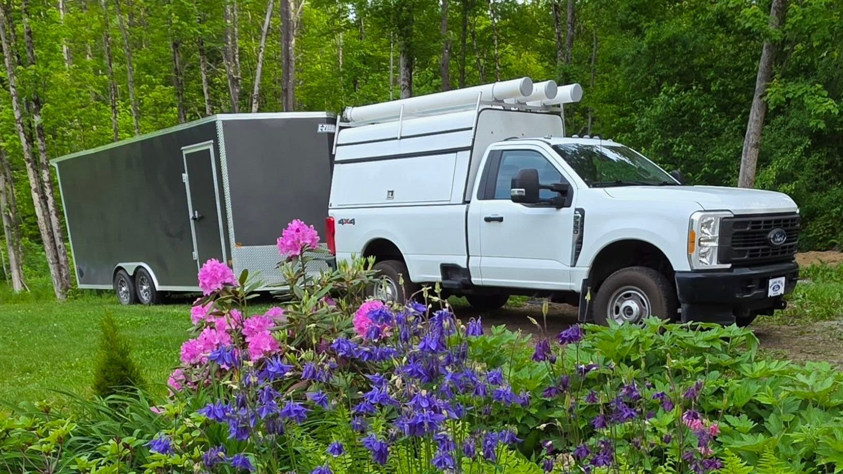 White pickup truck with enclosed trailer parked on a forest road, with purple flowers in the foreground