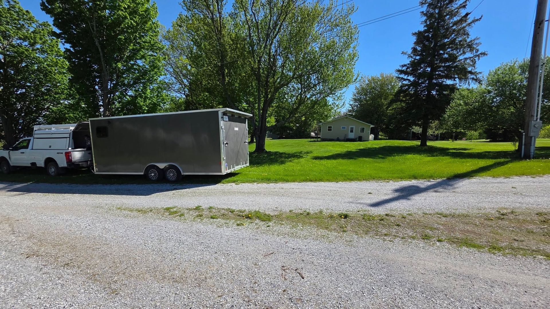 White truck towing a gray enclosed trailer on a gravel lot beside green trees and grass under a clear blue sky
