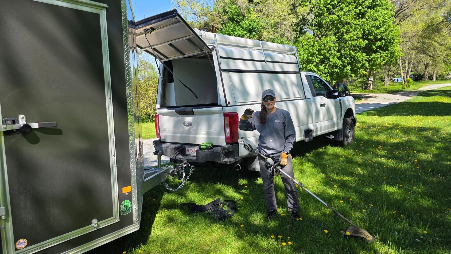 White pickup with canopy parked on grass, tailgate open, and a person standing beside it near a green trailer.