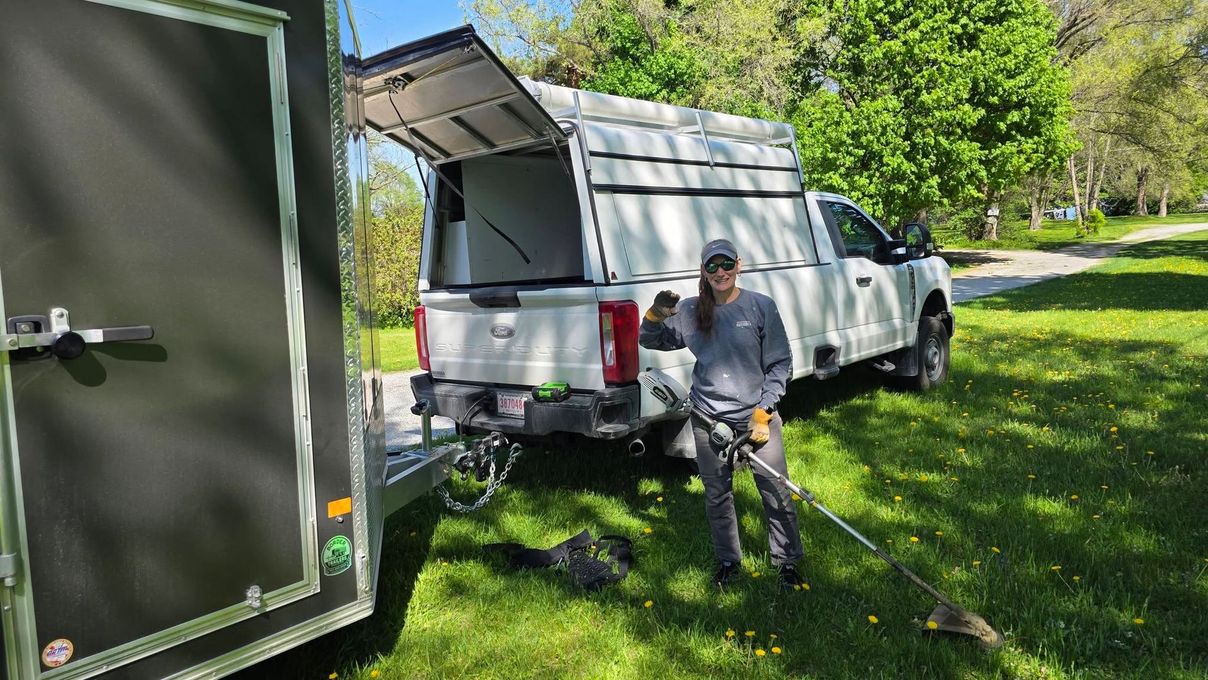 White pickup with canopy parked on grass, tailgate open, and a person standing beside it near a green trailer.