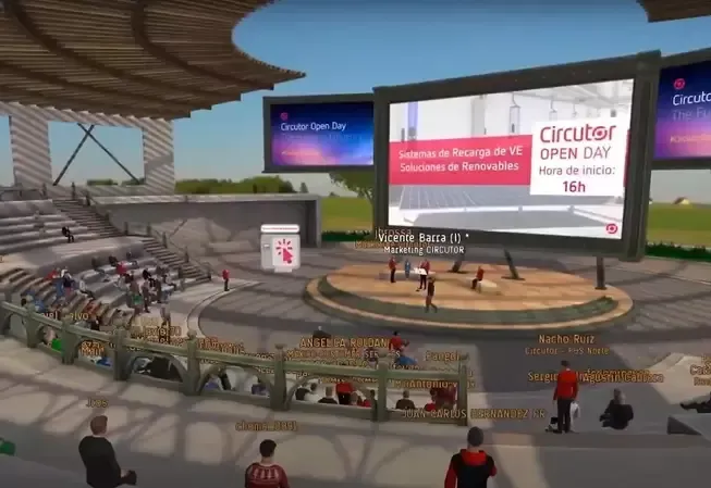 Virtual open day at a stadium-like venue. People on stage, large screen, audience in seats, sunny sky.