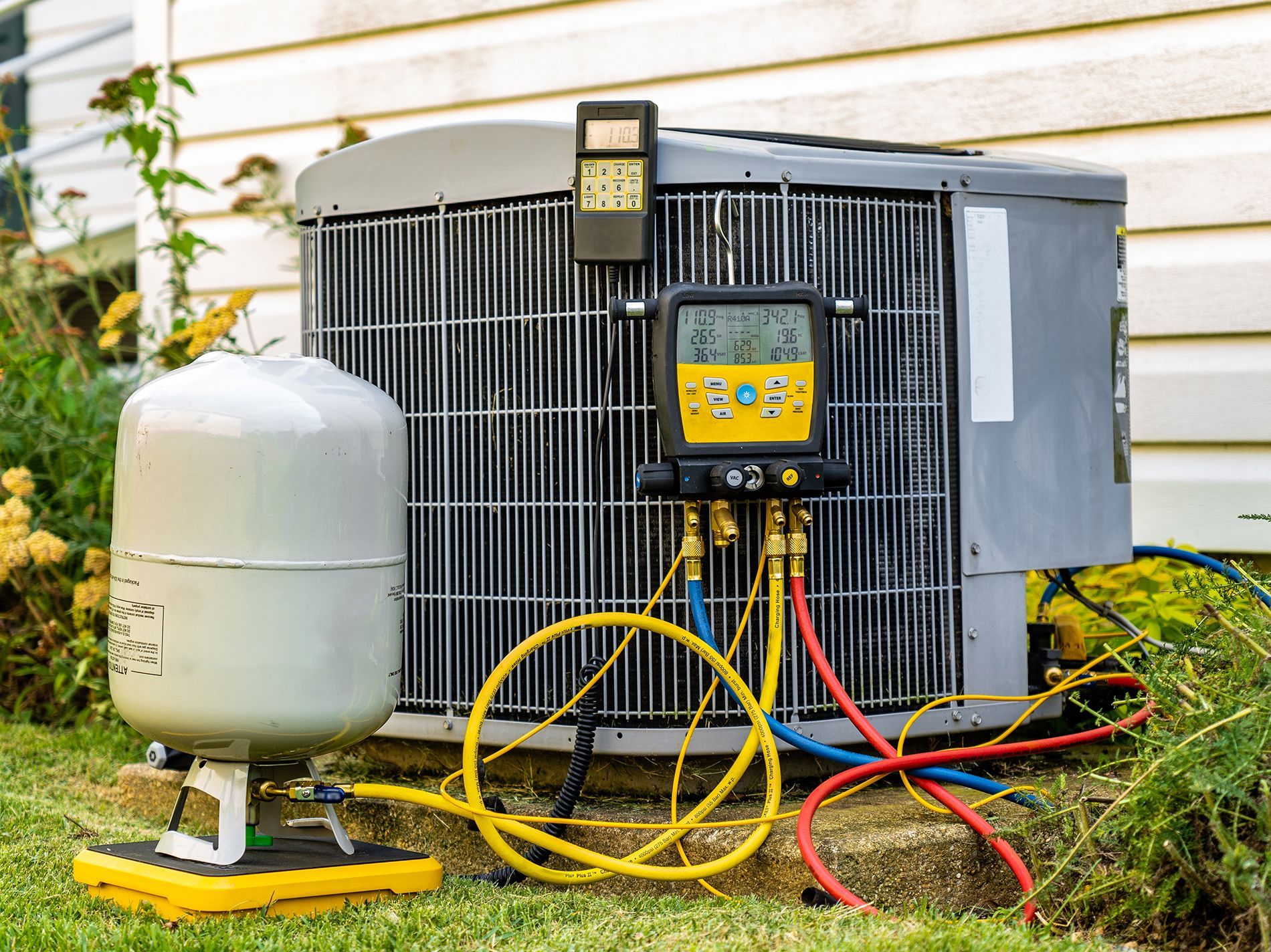 An HVAC technician's digital manifold gauge set connected to an outdoor air conditioning unit and a refrigerant tank.